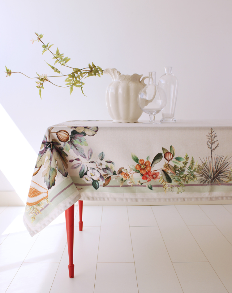 A tablecloth with a floral design, predominantly white with green and pink accents, displayed on a table along with a white vase with a floral design. New Classic; 150x250cm