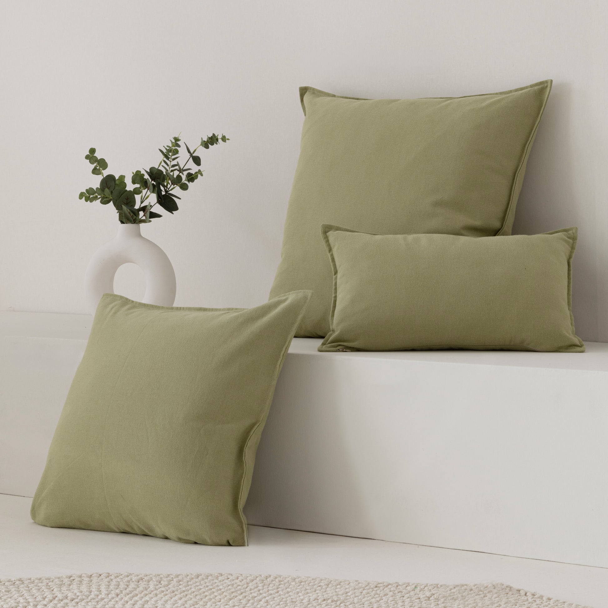 Three sage green throw pillows in different sizes arranged on a white bench against a white wall, with a small white vase of greenery beside them.
