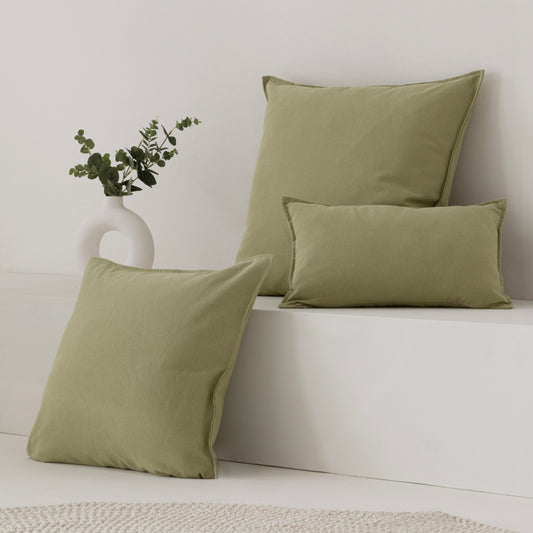 Three sage green throw pillows in different sizes arranged on a white bench against a white wall, with a small white vase of greenery beside them.