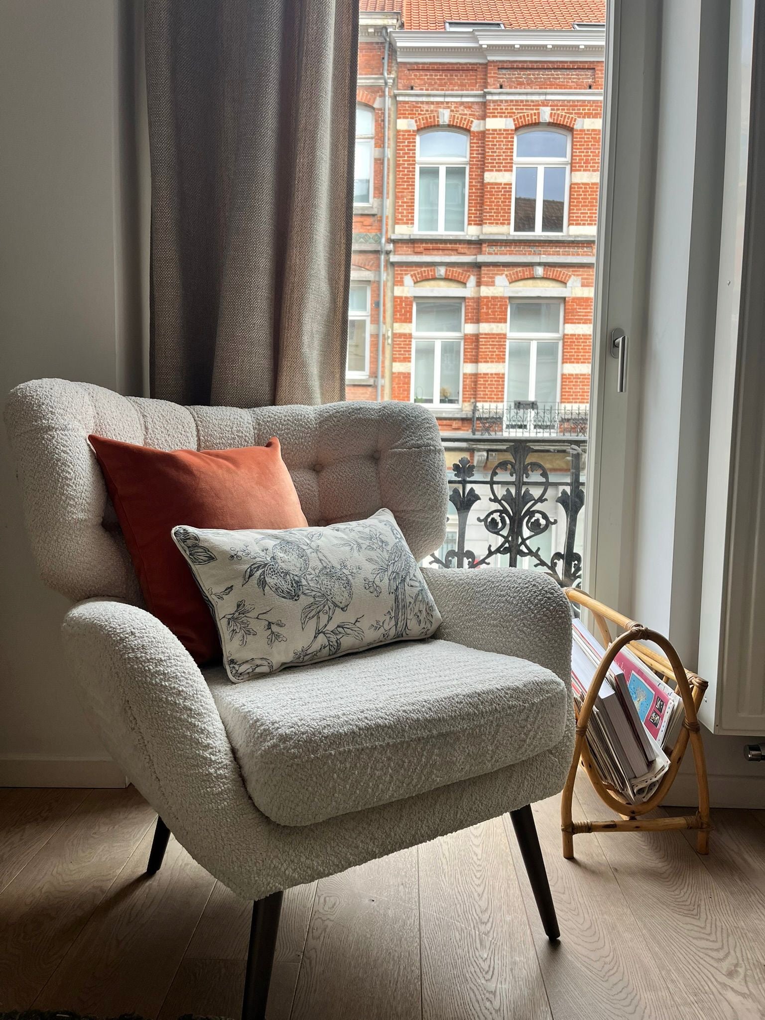 Armchair with orange and patterned cushions in a room with a window view of a brick building.