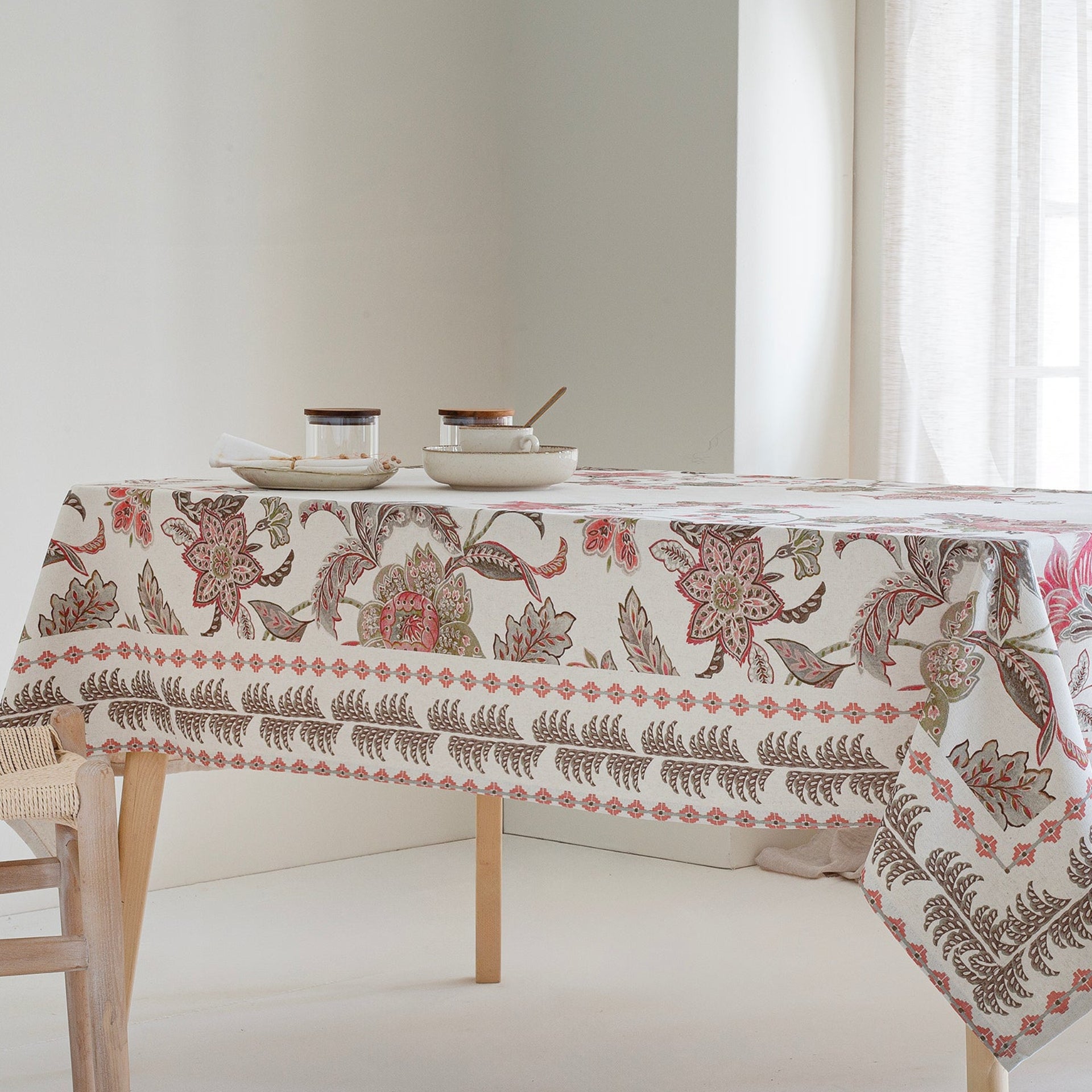 Table with a patterned tablecloth in a bright room with white curtains.
