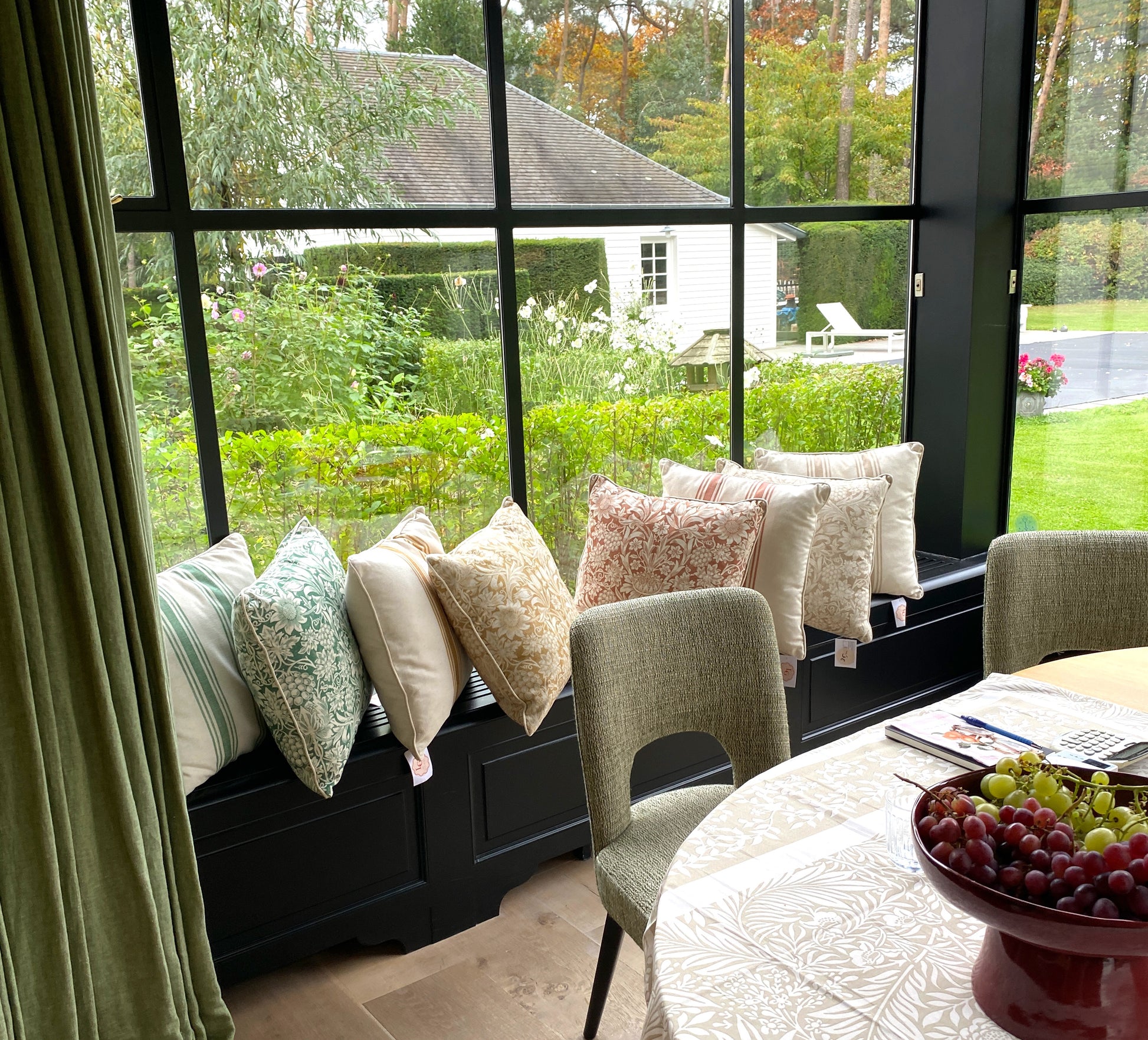Living room with a window seat filled with pillows with Wlliam Morris prints in red, yellow and green and a view of a garden. 