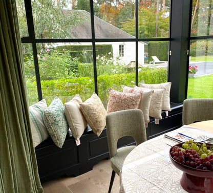Living room with a window seat filled with pillows with Wlliam Morris prints in red, yellow and green and a view of a garden. 