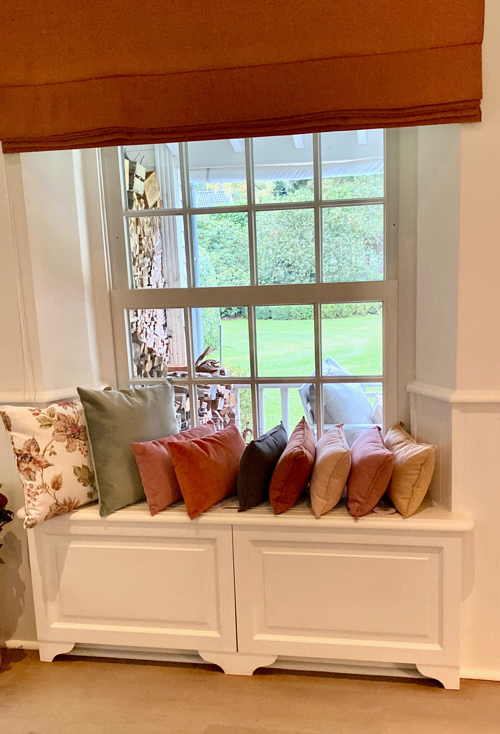 White bench with colorful pillows against a window with a terracotta curtain.