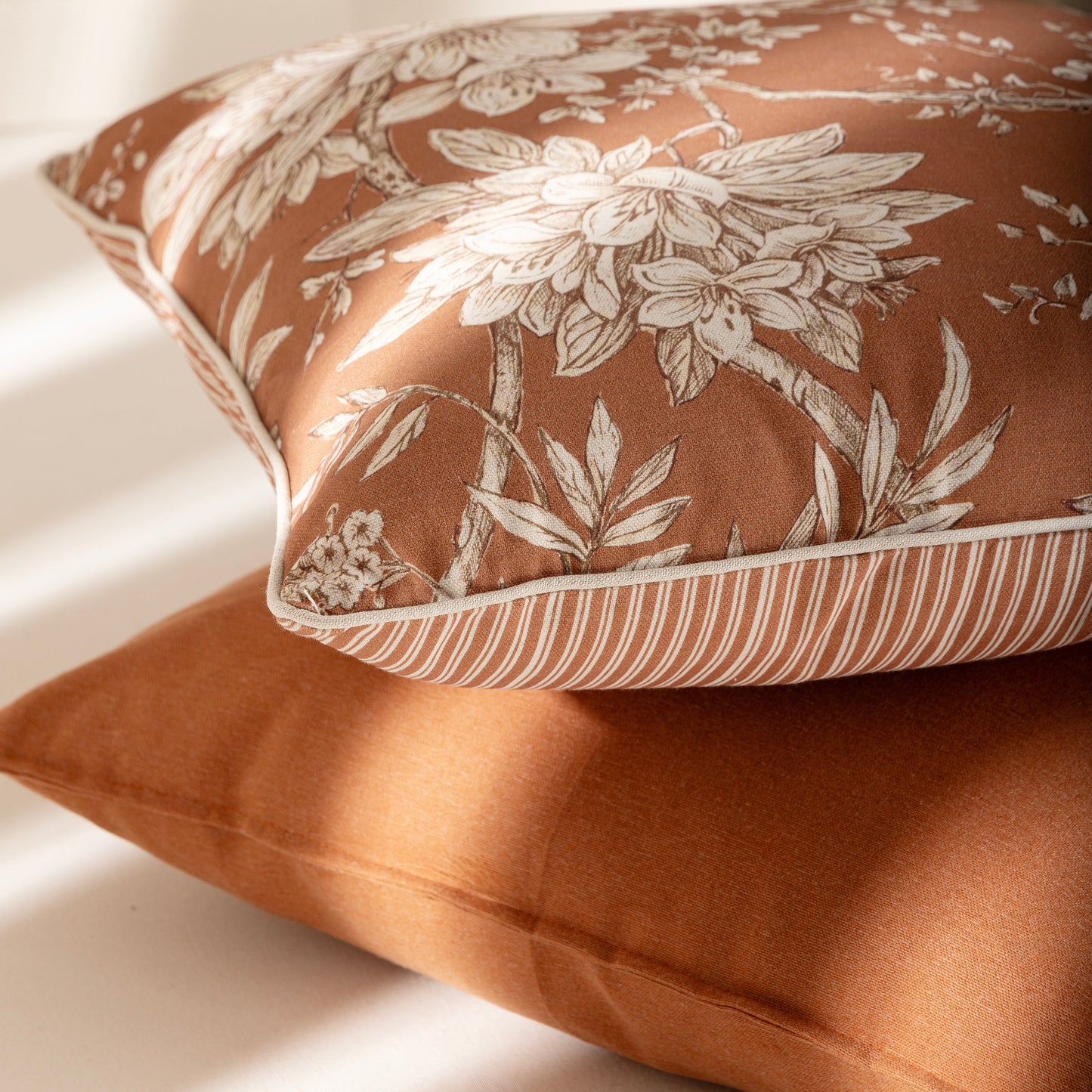 Two decorative throw pillows in warm rust and beige tones, with one pillow featuring a large floral toile pattern and the other solid, styled on a light surface with soft window shadows.