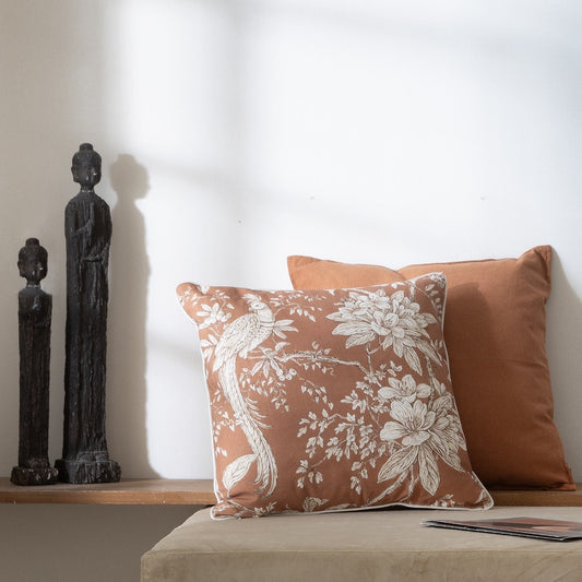 A decorative throw pillow with a white floral and bird print on a terracotta background sits in front of a solid terracotta pillow on a beige bench against a white wall.