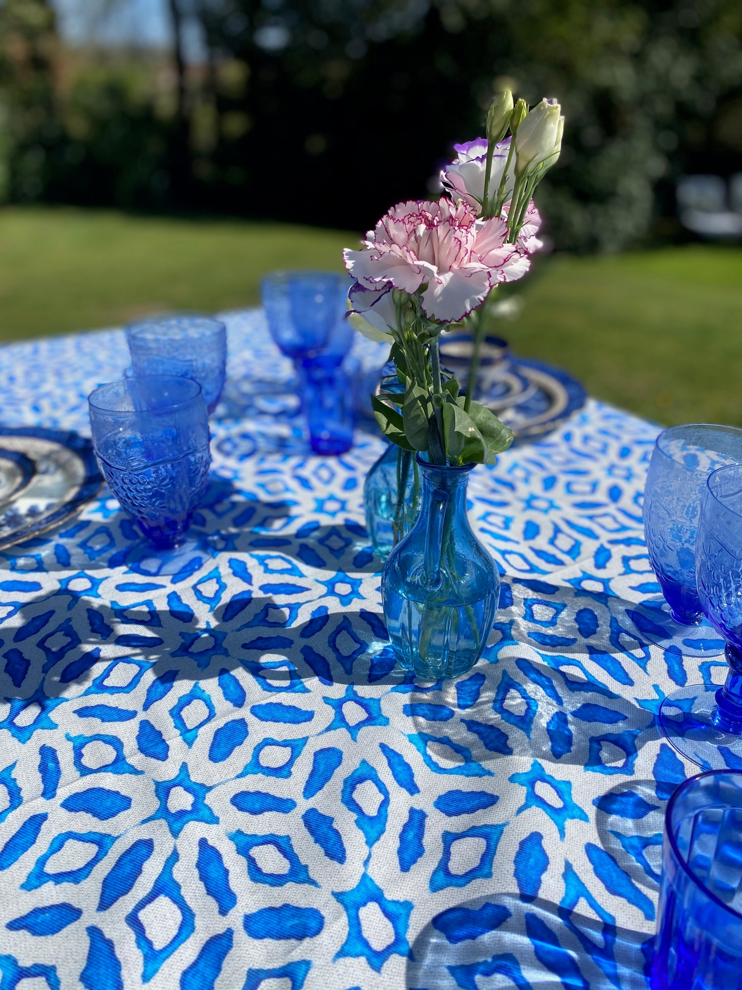 Table setting with blue glasses and a vase of pink flowers on a blue and white patterned tablecloth outdoors.