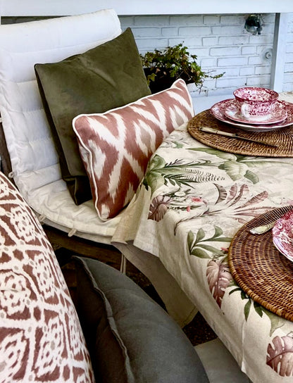 Decorative table setting with floral tablecloth, wicker chargers, and patterned pillows on a bench.