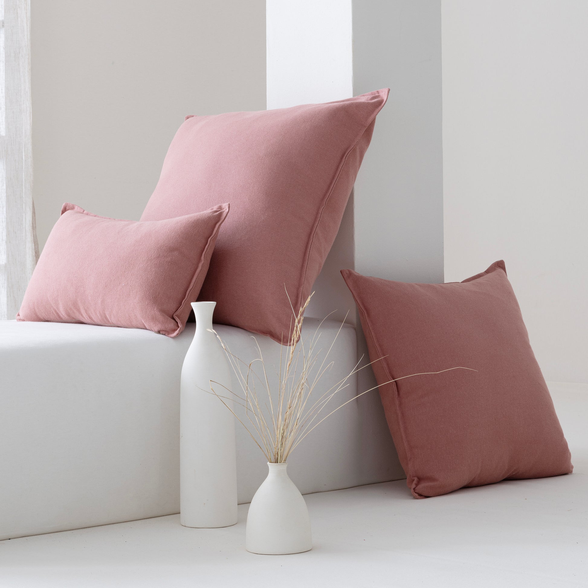 Three dusty rose throw pillows in different sizes styled on a white bench with two white ceramic vases and dried stems in a minimalist room setting.