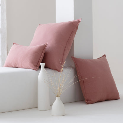 Three dusty rose throw pillows in different sizes styled on a white bench with two white ceramic vases and dried stems in a minimalist room setting.
